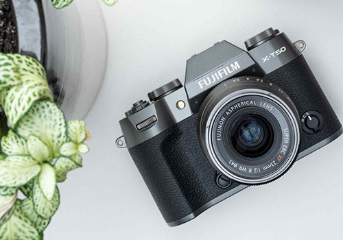 Fotocamera Fujifilm nera e grigia su un tavolo bianco, con una pianta in vaso sullo sfondo.