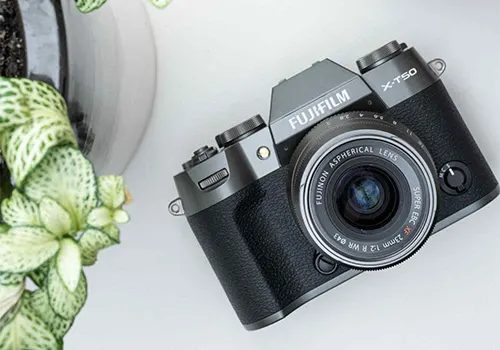 Fotocamera Fujifilm nera e grigia su un tavolo bianco, con una pianta in vaso sullo sfondo.