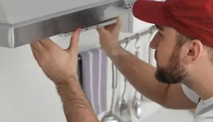 Un uomo con un berretto rosso installa una cappa aspirante in metallo in una cucina bianca, con utensili appesi sullo sfondo.