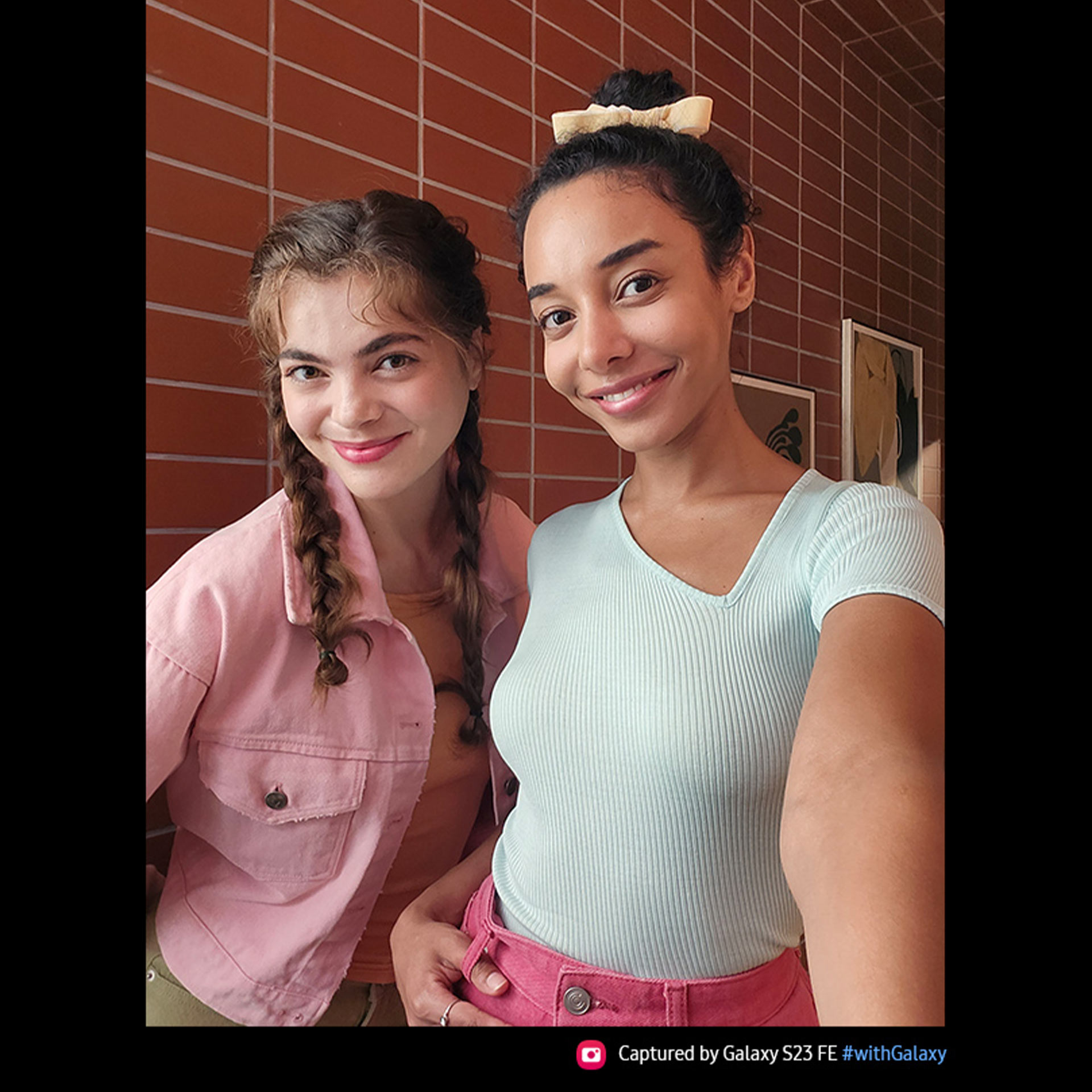 Due donne sorridono in primo piano, su sfondo di piastrelle rosse e cornici. Una indossa giacca rosa e trecce, l'altra maglia celeste e fiocco.