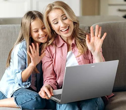 Madre e figlia sedute su un divano grigio salutano con la mano ad un laptop grigio.