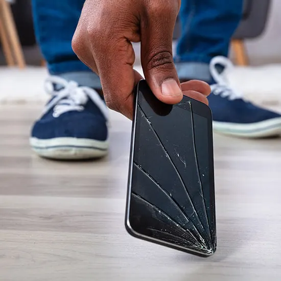 Una mano tiene un telefono rotto, con crepe sullo schermo. Sullo sfondo, scarpe blu e pavimento in legno chiaro.