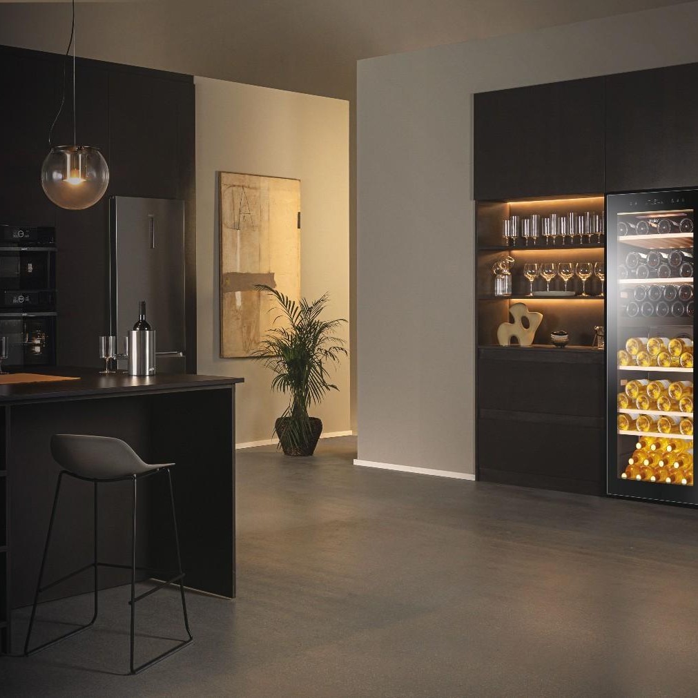 Interno di cucina con sgabello, bancone nero, frigo vini illuminato e scaffale con bicchieri. Quadro e pianta decorano.