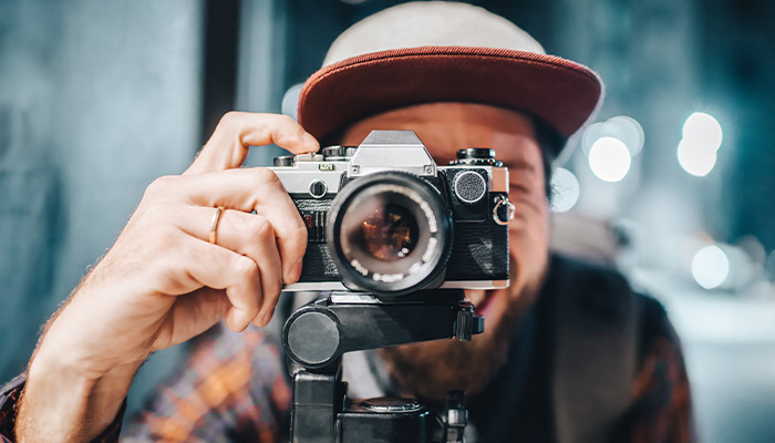 Un uomo con un cappello tiene una fotocamera vintage nera e argento su un treppiede, con sfondo sfocato.