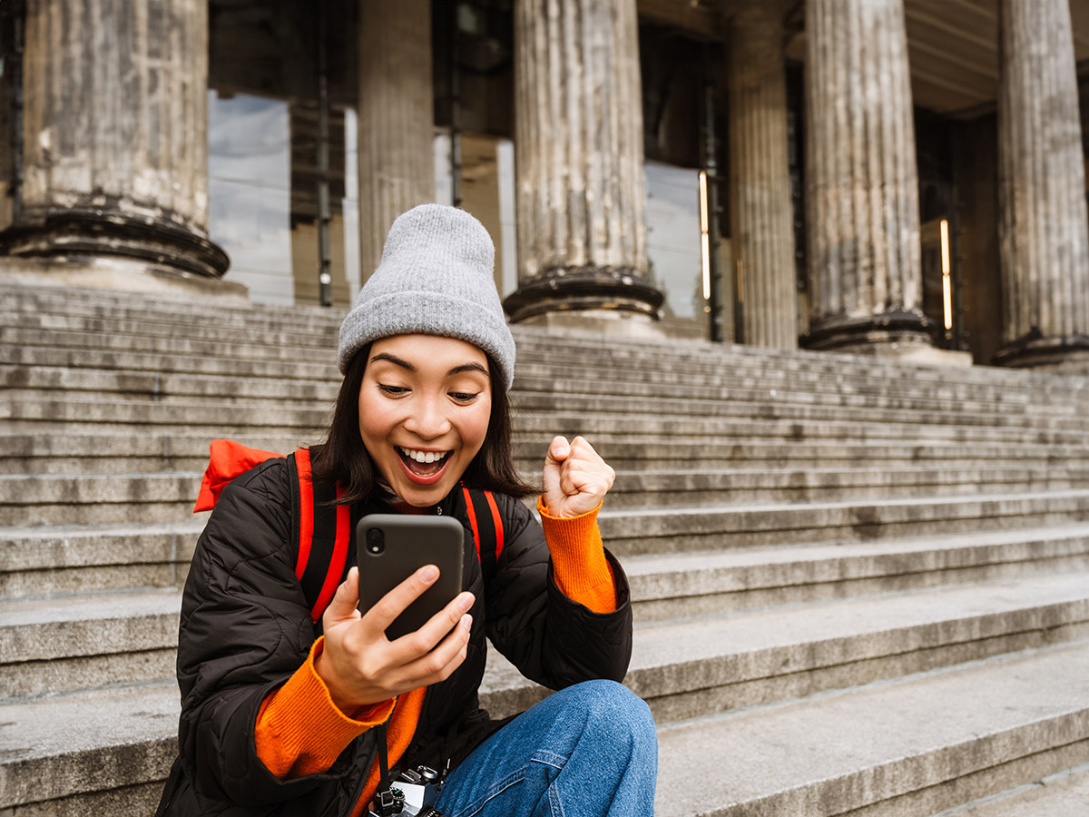 Donna seduta su scalinata di pietra con colonne sullo sfondo, esprime gioia guardando il telefono. Indossa giacca nera e berretto grigio.