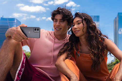 Un uomo e una donna sorridono mentre l'uomo tiene un telefono per fare un selfie. Sullo sfondo ci sono edifici alti e cielo blu.
