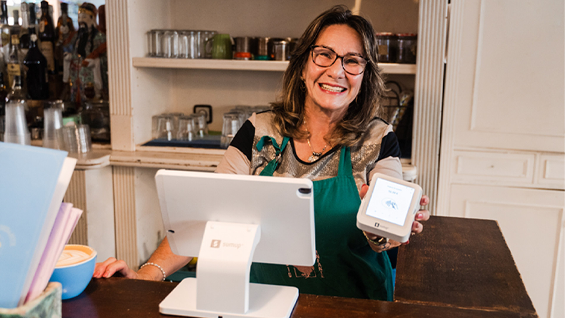 Donna sorridente con grembiule verde, mostra terminale di pagamento. Sul bancone, un tablet bianco e una tazza blu. Men� a sinistra.