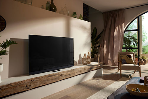 Soggiorno luminoso con TV nera su mensola in legno, poltrona beige e finestra ad arco. Elementi decorativi completano l'ambiente.