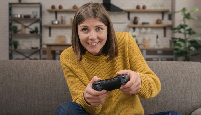 Una giovane donna sorride seduta sul divano mentre impugna un controller per videogiochi nero.