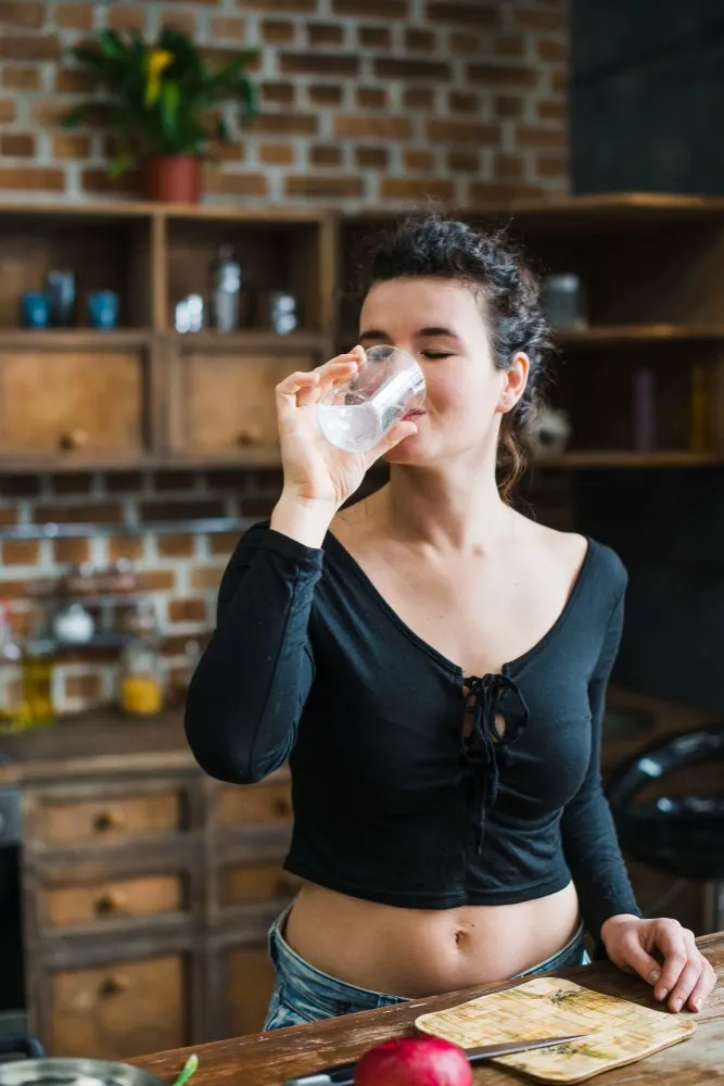 Donna beve acqua in cucina. Sul bancone ci sono un tagliere, un coltello e una melagrana rossa. Sullo sfondo scaffali e mattoni.