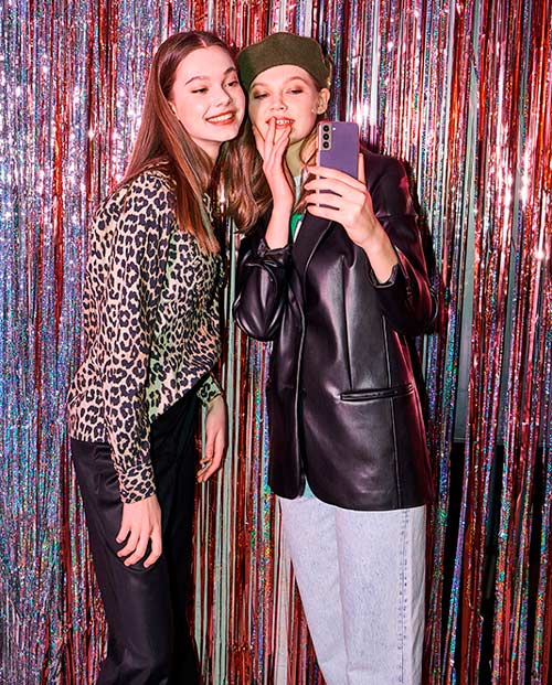 Due ragazze posano per una foto con un telefono viola davanti a uno sfondo di fili argentati e rossi. Una indossa una giacca leopardata.