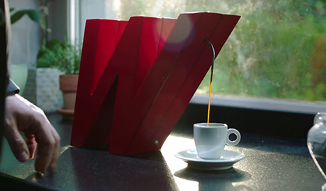 Moka rossa versa caff� in tazza bianca su piattino. Mano appoggiata al tavolo, finestra e piante sullo sfondo.