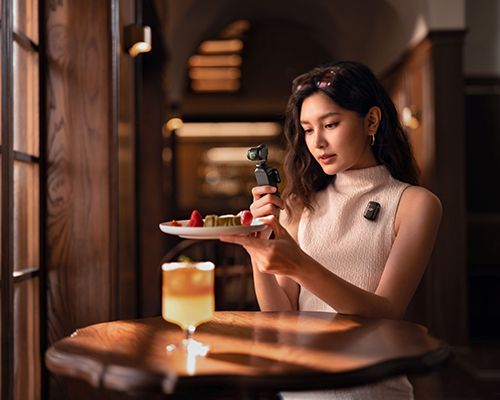 Donna riprende cibo con videocamera portatile. Tavolo con bevanda e finestra sullo sfondo.