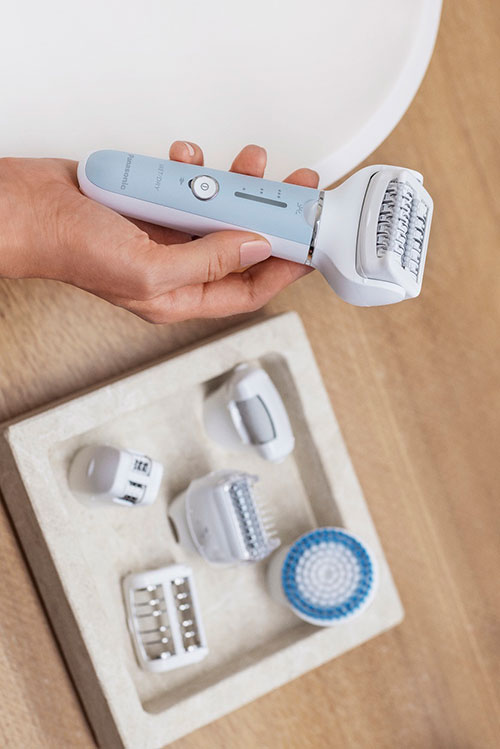 Mano che tiene un epilatore bianco e azzurro sopra una scatola con accessori su un tavolo di legno.