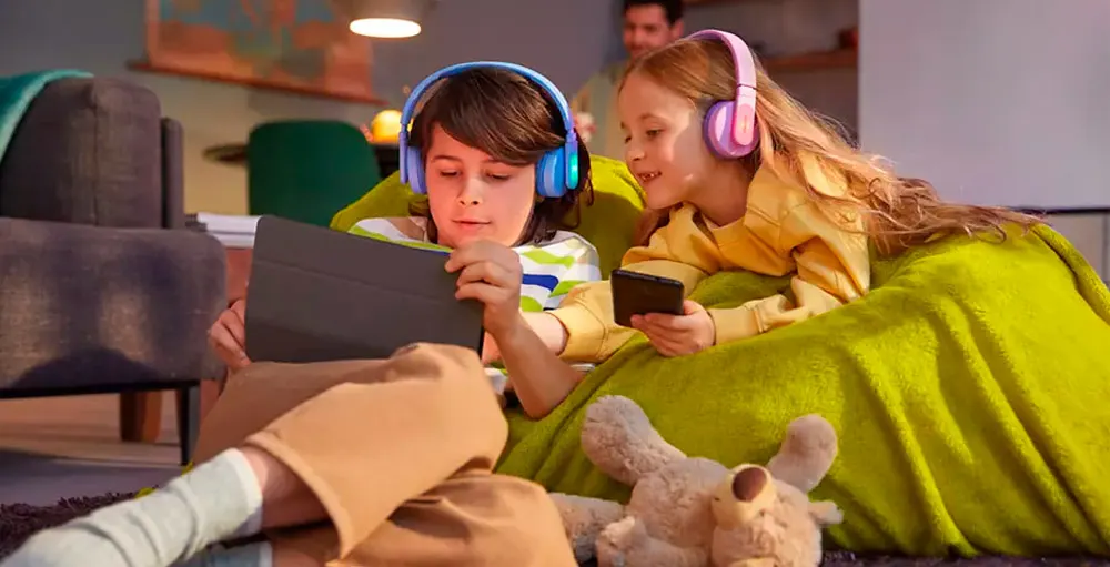 Due bambini con cuffie blu e rosa usano un tablet e un telefono su un pouf verde, con un orsacchiotto. Un adulto è sullo sfondo.