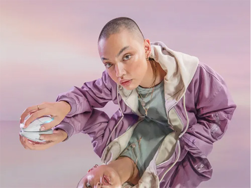 Donna con giacca viola e felpa grigia che tiene una confezione bianca su sfondo sfumato.