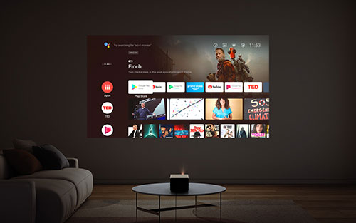 Soggiorno buio con divano grigio e tavolino rotondo. Schermo proiettato sulla parete mostra menu app di streaming.