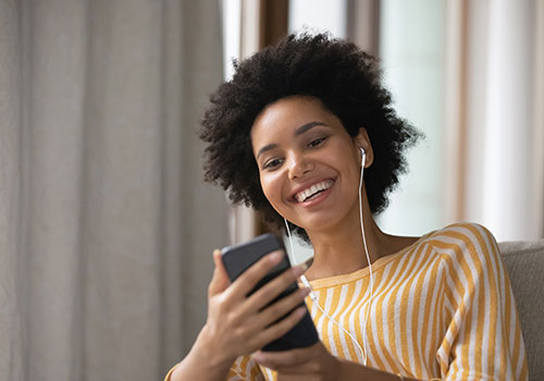 Donna sorridente con cuffie che tiene in mano un telefono. Indossa una camicia a righe gialle e bianche. Tenda sullo sfondo.