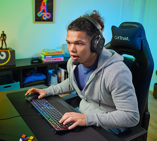 Un uomo con cuffie e microfono � seduto su una sedia da gaming nera, usando una tastiera multicolore. Sullo sfondo ci sono libri e decorazioni.