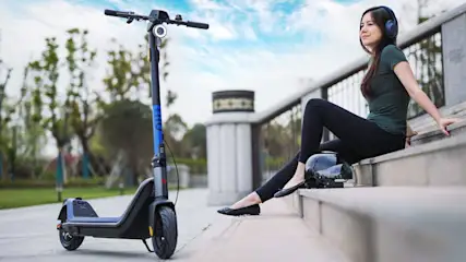 Donna seduta su gradini con cuffie, accanto a casco e monopattino elettrico su pavimentazione esterna. Alberi sullo sfondo.