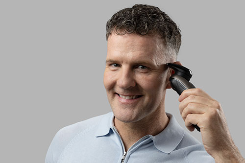 Uomo sorridente che usa un tagliacapelli nero vicino all'orecchio. Indossa una polo azzurra. Lo sfondo � grigio.