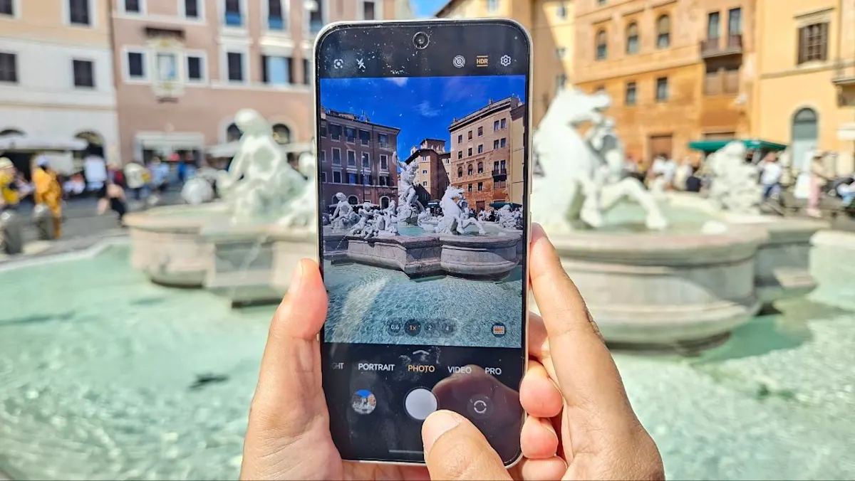 Mani tengono un telefono che inquadra una fontana con statue in una piazza affollata e palazzi sullo sfondo.