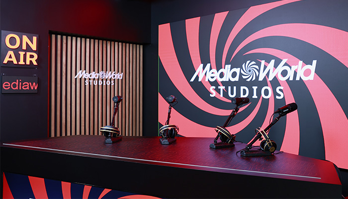 Studio con microfoni su tavolo nero, scritte "On Air" e "Media World Studios" su sfondo a spirale rossa e nera.