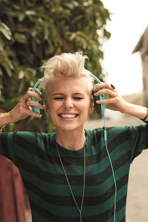 Donna sorride con cuffie azzurre, indossa maglione a righe verdi e nere e una collana dorata. Sfocato sfondo urbano e vegetazione.