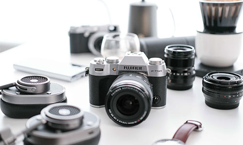 Fotocamera Fujifilm nera e argento circondata da obiettivi, cuffie, un orologio e accessori da caff�.