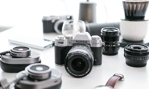 Fotocamera Fujifilm nera e argento circondata da obiettivi, cuffie, un orologio e accessori da caff�.