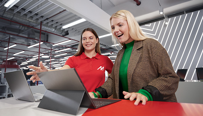 Due donne guardano dei laptop su un tavolo rosso. Una indossa una polo rossa con una "M". Dietro, luci e un soffitto industriale.
