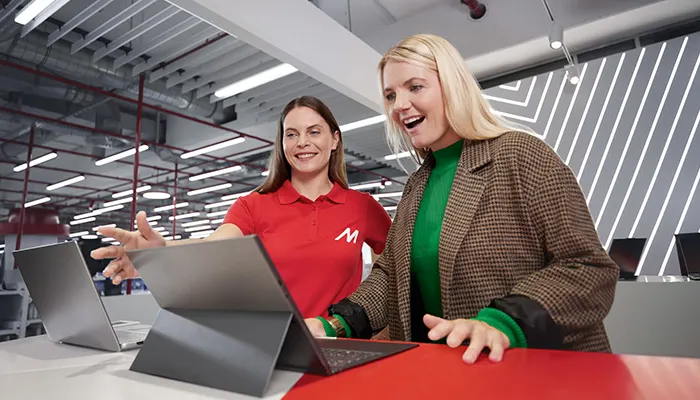 Due donne guardano dei laptop su un tavolo rosso. Una indossa una polo rossa con una "M". Dietro, luci e un soffitto industriale.