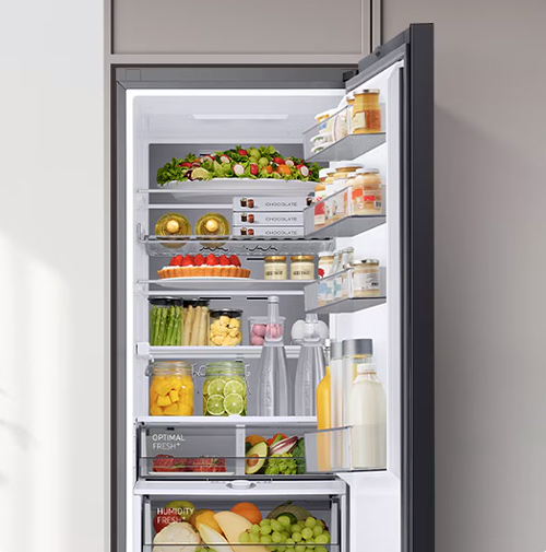 Frigorifero aperto pieno di alimenti, inclusi insalata, frutta, verdura, salse e bevande, su diversi ripiani e cassetti.