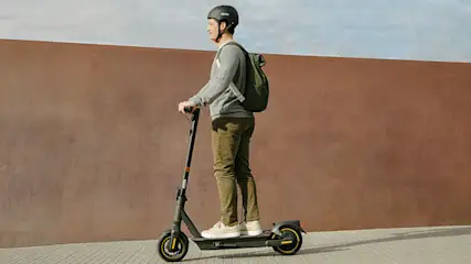 Un uomo con casco e zaino guida un monopattino elettrico su un marciapiede. Sullo sfondo, un muro marrone chiaro e il cielo.
