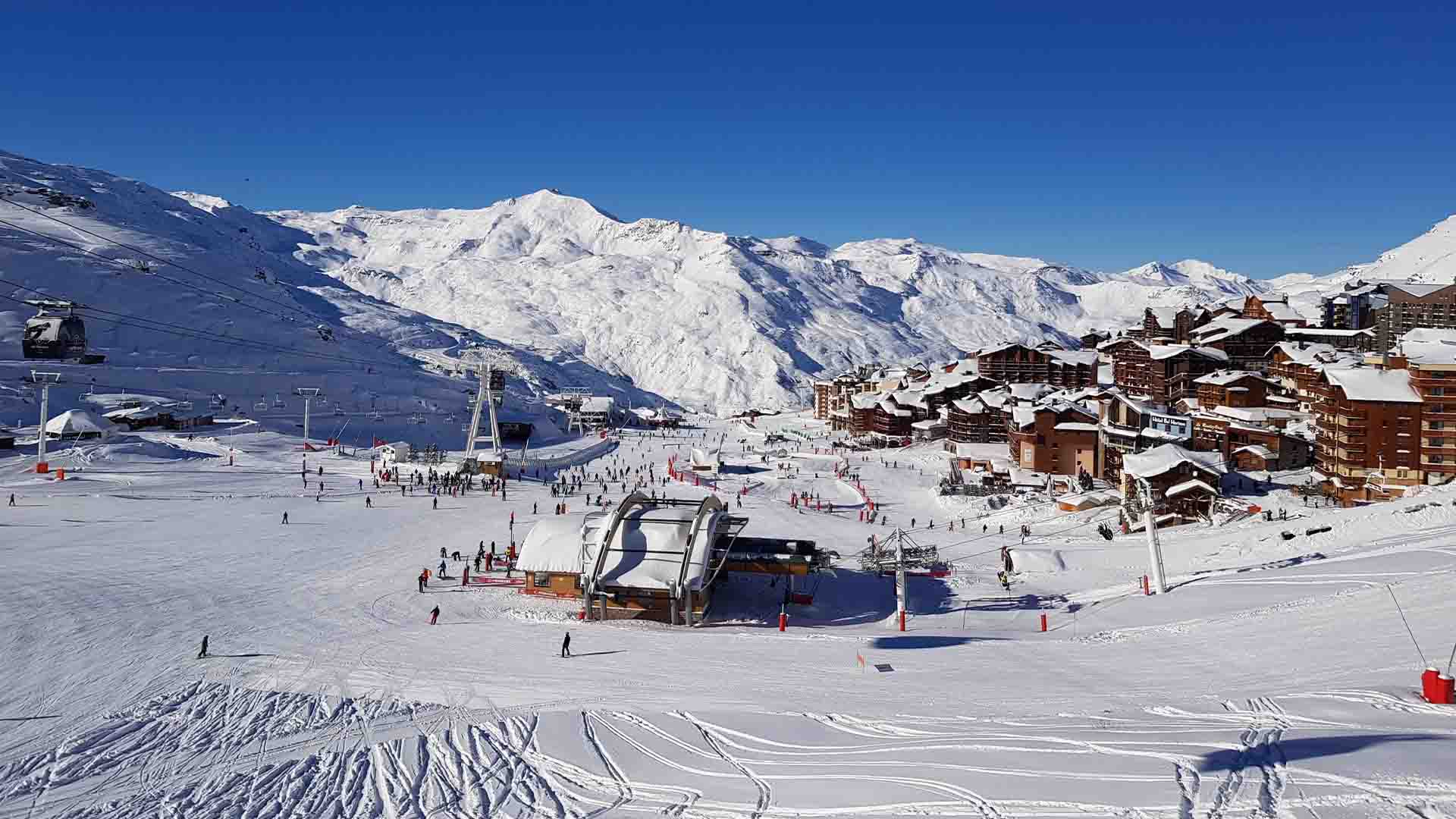 Paesaggio montano innevato con impianti di risalita, persone che sciano e edifici residenziali. Cielo azzurro limpido.