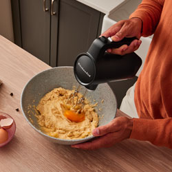 Ciotola con impasto e uovo, persona usa sbattitore elettrico. Mobile grigio sullo sfondo.