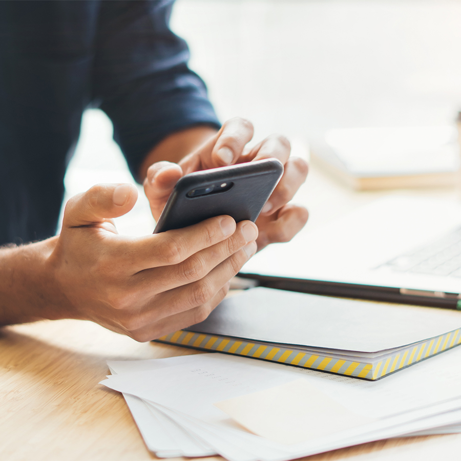 Uomo con camicia scura usa un telefono. Sul tavolo ci sono un quaderno con dettagli gialli, fogli di carta e una penna.