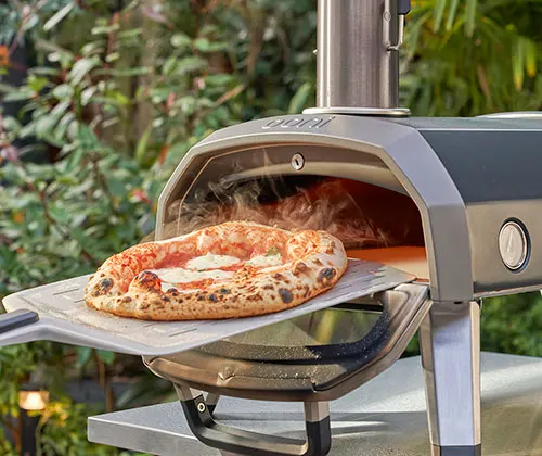 Pizza su pala di metallo davanti a forno da esterno grigio. Sullo sfondo, vegetazione verde.