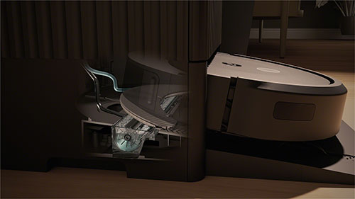 Un aspirapolvere robot bianco e marrone è parcheggiato su una base scura vicino a un mobile scuro, su un pavimento chiaro.