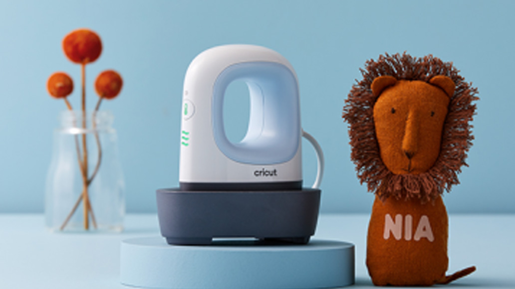 Su uno sfondo blu, un leone di peluche con la scritta "NIA", un ferro da stiro Cricut e un vaso con fiori arancioni.