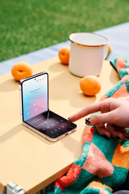 Su un tavolo, un telefono aperto mostra un'app musicale. Vicino, albicocche, una tazza bianca e una coperta colorata. Una mano tocca lo schermo.