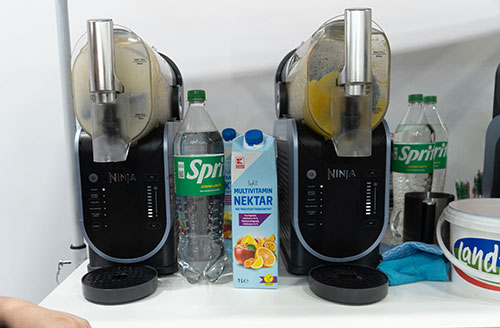 Due frullatori Ninja neri su un tavolo bianco, con bevande, una bottiglia di Sprite e un cartone di succo di frutta.