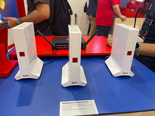 Tre router bianchi con antenne su tavolo blu e rosso. Sullo sfondo, persone in piedi e cartello con testo.
