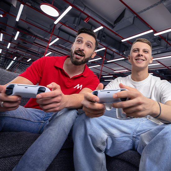 Due uomini seduti su un divano grigio, giocano con controller bianchi in un ambiente illuminato da luci al neon.