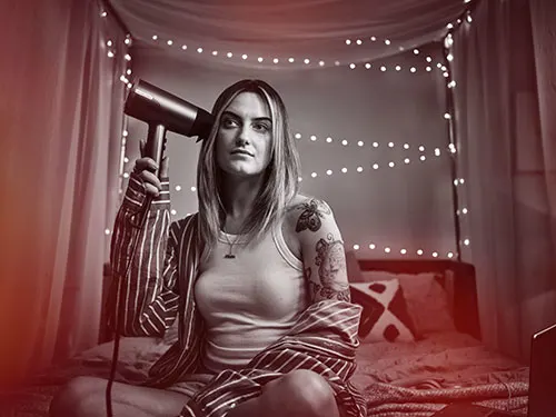 Donna con asciugacapelli in camera da letto con luci. Indossa canottiera, giacca a righe, ha tatuaggi sul braccio.