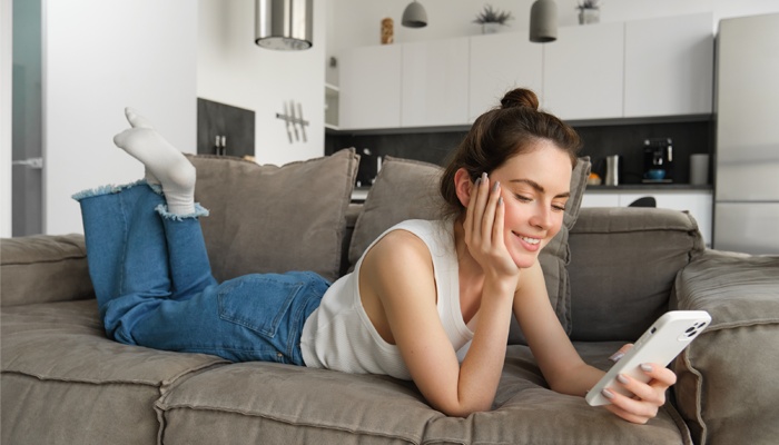 Una donna sorridente è sdraiata a pancia in giù su un divano grigio mentre guarda lo smartphone, con una cucina moderna sullo sfondo.