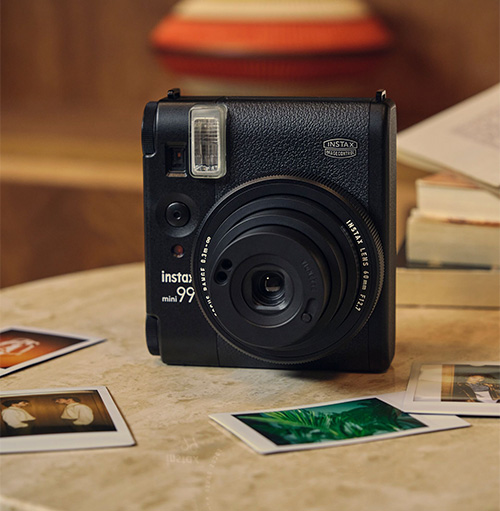 Fotocamera Instax nera su un tavolo beige con foto polaroid. Sullo sfondo si vedono libri e un oggetto decorativo.