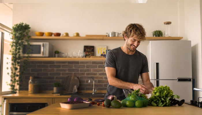 Un uomo sorridente prepara della verdura fresca sull'isola di una cucina moderna e luminosa, con un frigorifero e un forno a microonde sullo sfondo.