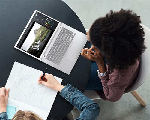 Vista dall'alto di un tavolo con un laptop aperto, una persona che scrive su carta e un'altra seduta.