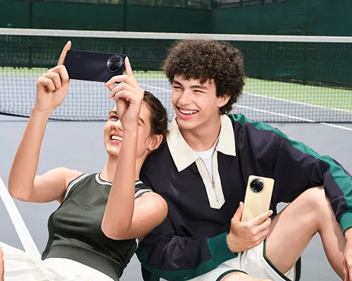 Due persone sorridenti con telefoni su un campo da tennis. Una persona scatta una foto, l'altra tiene il telefono. Rete da tennis sullo sfondo.